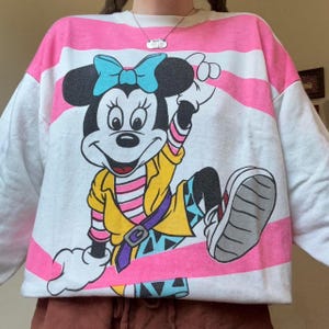 May include: White sweatshirt with pink stripes and a large graphic of Minnie Mouse. Minnie is wearing a blue bow, yellow jacket, and colorful patterned pants. The sweatshirt has long sleeves.