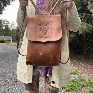 May include: A tan leather backpack featuring a rectangular flap with a debossed pinecone design. The bag has two thin shoulder straps and a vertical rectangular body with visible natural leather grain and light scuff marks on the surface.