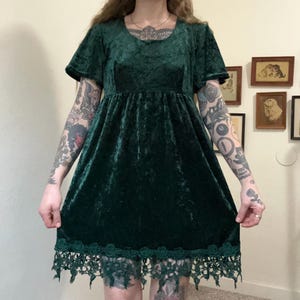 May include: A dark green velvet dress with short sleeves and a scoop neckline. The dress has a fitted bodice and a flared skirt, with a decorative lace trim at the hem. The dress is a mini dress.