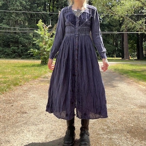 May include: A long-sleeved, button-down midi dress in a deep purple hue. The bodice features intricate dark purple embroidery and a smocked waistline. The skirt is pleated and flows to a mid-calf length, complemented by black leather ankle boots.