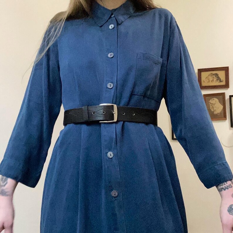 Puede incluir: Un vestido camisa de manga larga de color azul oscuro con cierre de botones en la parte delantera y un solo bolsillo en el pecho izquierdo. El vestido est&aacute; ce&ntilde;ido a la cintura con un cintur&oacute;n de cuero negro.