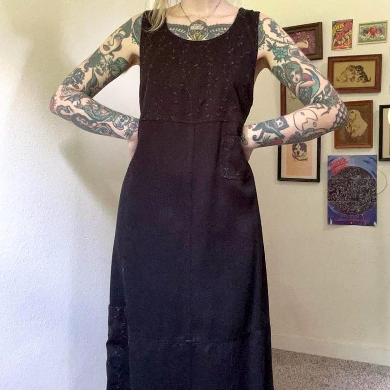 May include: A sleeveless black midi dress featuring a scoop neckline and a subtle tonal textured pattern on the bodice and a small patch pocket on the chest. The dress has a straight silhouette with a horizontal seam detail across the lower skirt.