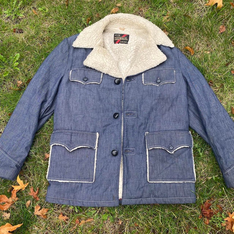 May include: A blue denim jacket with a white faux fur collar and two large pockets. The jacket has a button closure and a white stitching detail around the pockets.