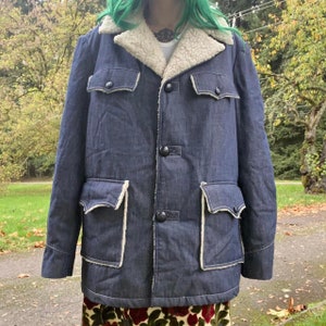 May include: A blue denim jacket with a white faux fur collar and lining. The jacket has two large flap pockets on the front and a button closure.