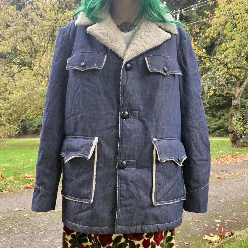 May include: A blue denim jacket with a white faux fur collar and lining. The jacket has two large flap pockets on the front and a button closure.
