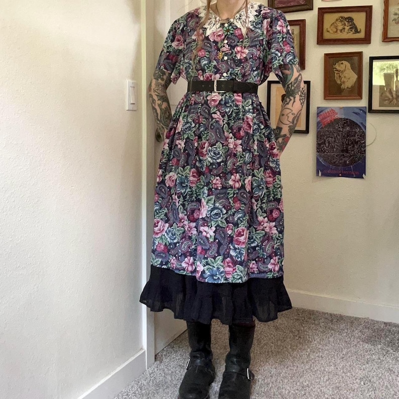 Puede incluir: Un vestido midi con estampado floral y de cachemira multicolor sobre fondo azul oscuro, con cuello de encaje blanco, cierre frontal de botones, cintur&oacute;n negro en la cintura y bajo con volantes negros, combinado con botas de cuero negro.