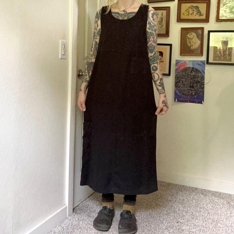 May include: A sleeveless, ankle-length black dress featuring a scoop neckline and a relaxed, straight-cut silhouette. The garment is made of a solid black fabric and includes a visible patch pocket on the lower right side of the skirt area.