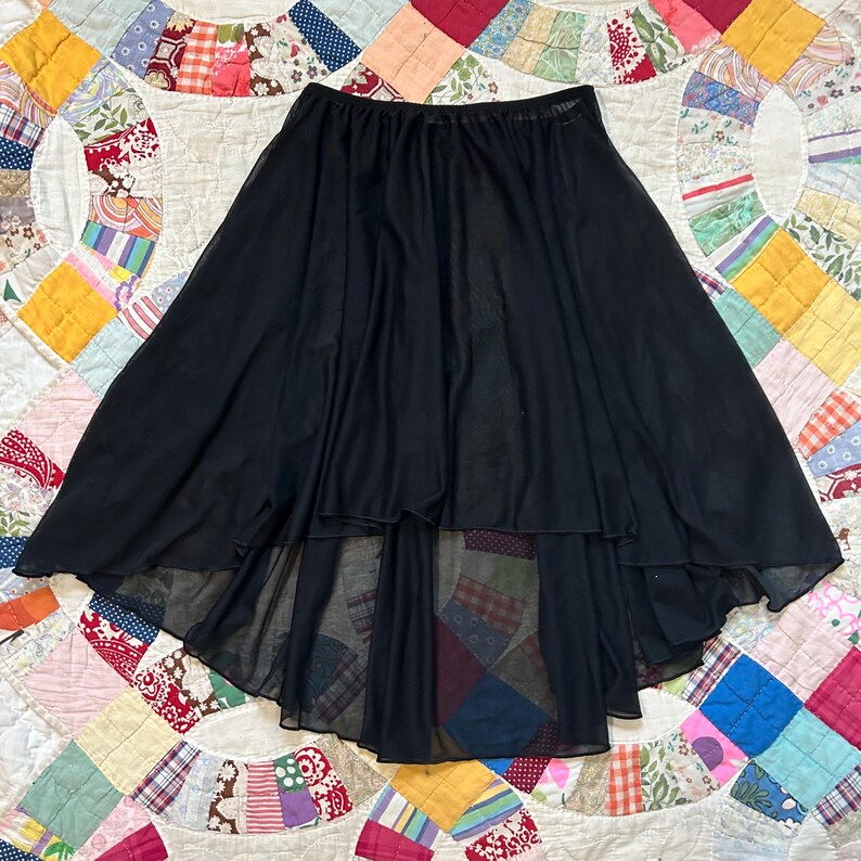 May include: A black, high-low skirt with a flowing, layered design. The skirt has a wide elastic waistband and a sheer, lightweight fabric. The skirt's hemline is shorter in the front and longer in the back.