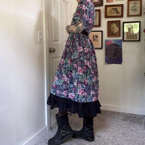 Puede incluir: Un vestido midi con base azul oscuro y un estampado floral y de cachemira en tonos rosa, morado y azul verdoso. El vestido tiene mangas cortas, una cintura ajustada con un cintur&oacute;n negro y un dobladillo negro con volantes en la parte inferior.