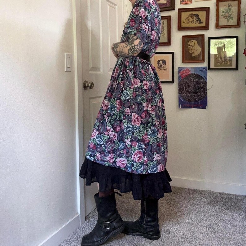 Puede incluir: Un vestido midi con base azul oscuro y un estampado floral y de cachemira en tonos rosa, morado y azul verdoso. El vestido tiene mangas cortas, una cintura ajustada con un cintur&oacute;n negro y un dobladillo negro con volantes en la parte inferior.