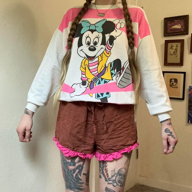 May include: A white and pink striped sweatshirt featuring a large graphic of Minnie Mouse. The cartoon character is wearing a yellow jacket and a blue bow. Paired with brown shorts with a pink ruffle trim.