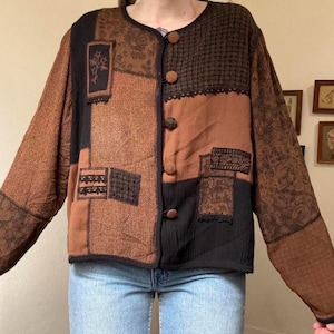 May include: A patchwork jacket in shades of brown and black. The jacket features a round neckline, large wooden buttons, and long sleeves. The design incorporates various textured fabrics and decorative elements, creating a unique and artistic look. The jacket is worn over blue jeans.