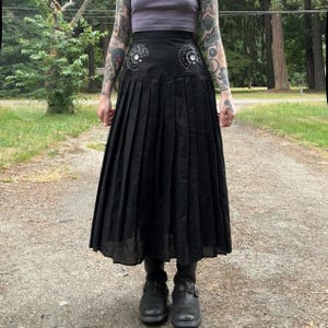May include: A black midi-length pleated skirt featuring a high-waisted design with two decorative semi-circular patches on the front waistband. Each patch is adorned with white stitching and a single central button detail, creating a distinct look.