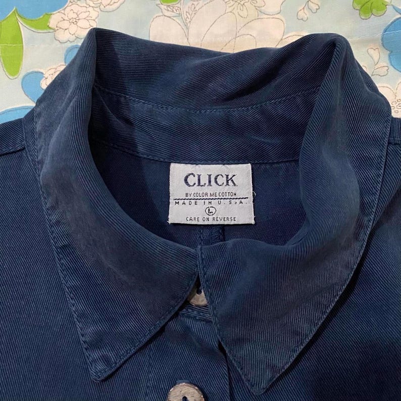 Puede incluir: Una camisa vaquera azul oscuro con una etiqueta blanca que dice "CLICK BY COLOR ME COTTON MADE IN U.S.A. L CARE ON REVERSE".