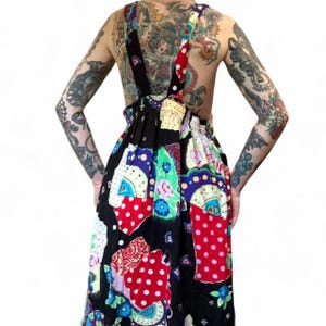 May include: A sleeveless jumper dress featuring a vibrant patchwork design with a black base. The pattern includes various colorful sections with red and white polka dots, floral motifs, and abstract shapes. It has wide shoulder straps and a gathered waist.