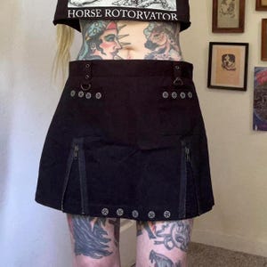 May include: Black mini skirt with silver hardware, zippers, and decorative rivets. The skirt has a structured design with a slight A-line shape. The top of the image shows a black crop top with the text "HORSE ROTOVATOR".