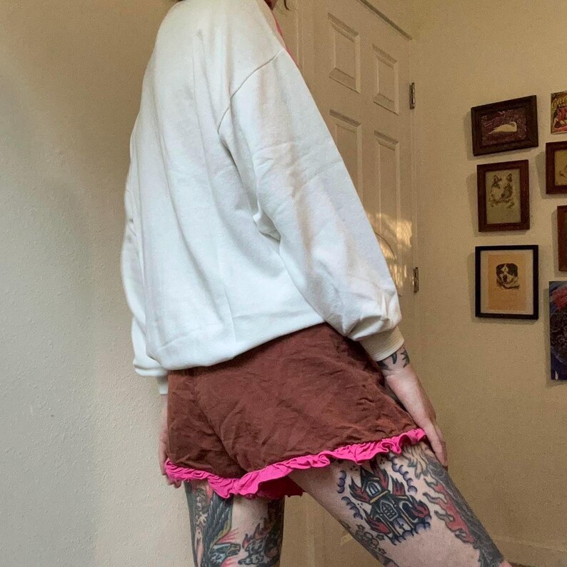 May include: A white oversized sweatshirt paired with brown shorts featuring a pink ruffle trim. The casual outfit is captured from a side angle, showcasing the relaxed fit of the clothing. The shorts are a dark brown color.