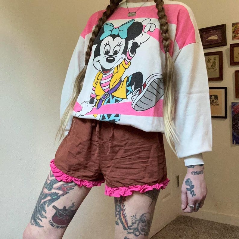 May include: A white and pink striped sweatshirt featuring a Minnie Mouse graphic, paired with brown shorts trimmed with pink ruffles. The outfit is casual and vintage-inspired, suitable for everyday wear.