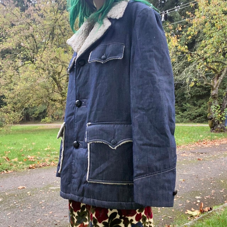 May include: A dark blue denim jacket with a sherpa collar and large patch pockets. The jacket has a button closure and a contrasting white stitching detail.