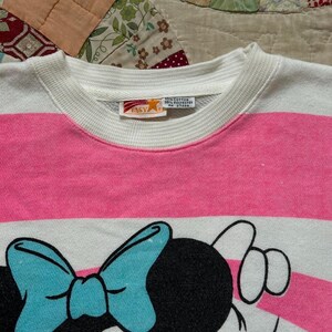 May include: A white and pink striped sweatshirt featuring a cartoon Minnie Mouse design. The design includes a large blue bow and a black head. The sweatshirt has a white collar and cuffs.