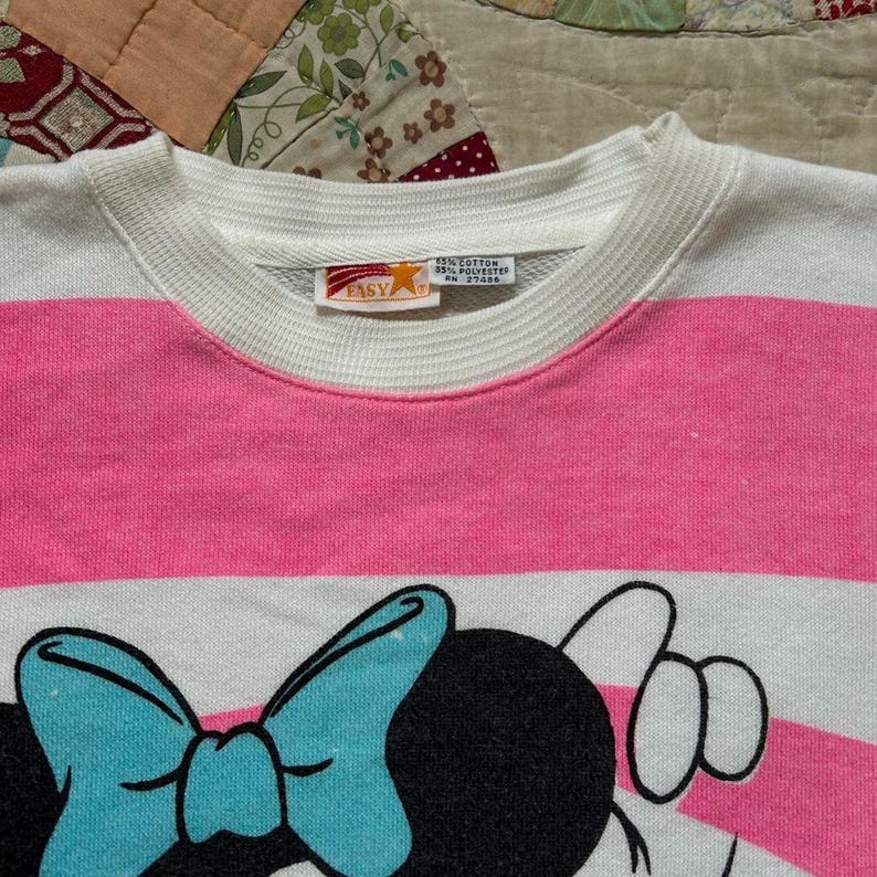 May include: A white and pink striped sweatshirt featuring a cartoon Minnie Mouse design. The design includes a large blue bow and a black head. The sweatshirt has a white collar and cuffs.