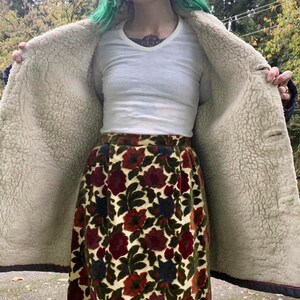 May include: A woman wearing a white shirt and a brown and blue floral skirt. She is wearing a brown jacket with a white fleece lining. The jacket is open and the fleece lining is visible.