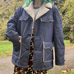 May include: A dark blue denim jacket with a white sherpa lining. The jacket has a button closure, two large pockets on the front, and a smaller pocket on the left chest. The jacket is lined with a white sherpa fabric.