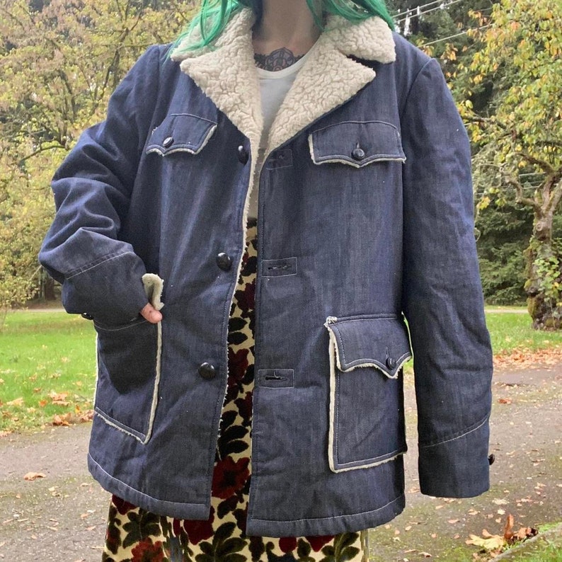 May include: A dark blue denim jacket with a white sherpa lining. The jacket has a button closure, two large pockets on the front, and a smaller pocket on the left chest. The jacket is lined with a white sherpa fabric.