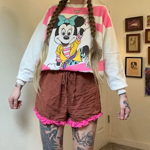 May include: A white and pink striped sweatshirt featuring a Minnie Mouse graphic. The sweatshirt is paired with brown shorts with a pink ruffle trim. The outfit is casual and colorful, suitable for everyday wear.