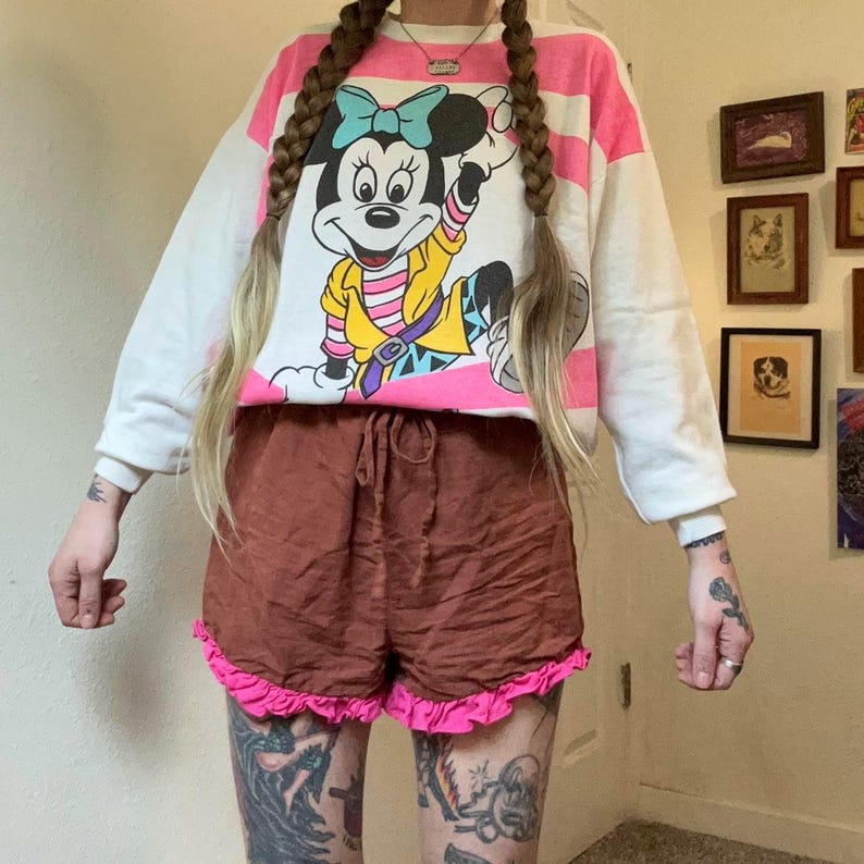May include: A white and pink striped sweatshirt featuring a Minnie Mouse graphic. The sweatshirt is paired with brown shorts with a pink ruffle trim. The outfit is casual and colorful, suitable for everyday wear.