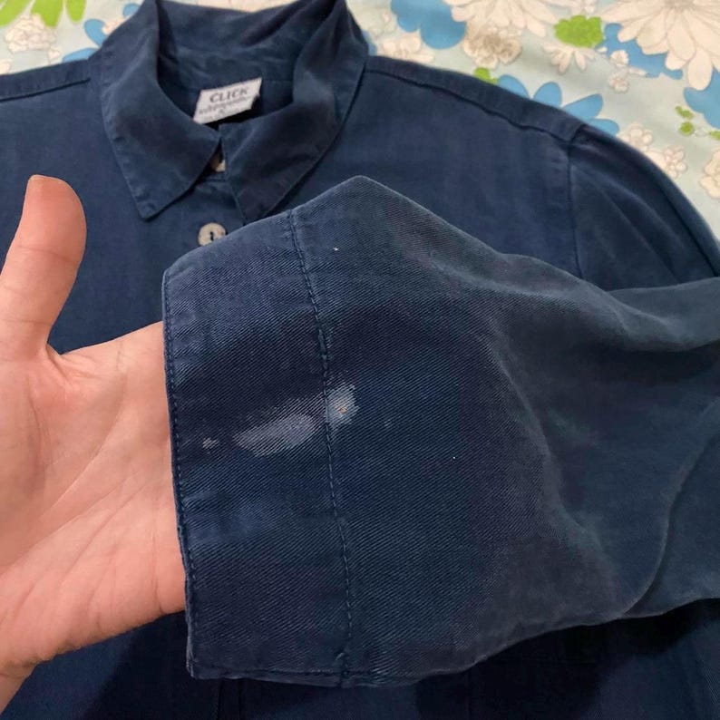 Puede incluir: Una camisa de manga larga azul oscuro con una etiqueta blanca que dice "Click" en el cuello. La camisa tiene un cierre de botones en la parte delantera y un solo bot&oacute;n en el pu&ntilde;o.