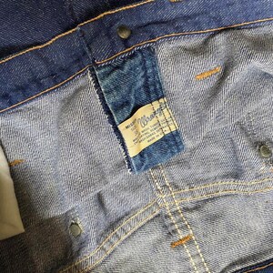 Puede incluir: Primer plano de un bolsillo de mezclilla azul con una etiqueta Wrangler cosida. La etiqueta es blanca con texto negro que dice "Misses Size 32" y "Wash, Tumble Dry, Remove Promptly, Machine Wash, Dark Separately, Made in USA".
