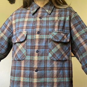 May include: A button-down plaid shirt in shades of blue, brown, and teal. The shirt has two front pockets and a collared neckline. The fabric appears to be a soft, woven material.