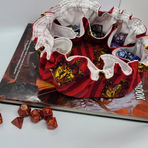 May include: Red and gold fabric dice bag with a Hogwarts house crest pattern. The bag is filled with various colored dice and is sitting on top of a book.