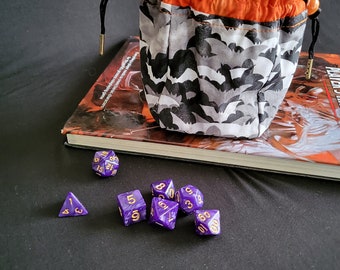 May include: A purple and gold dice bag with a black drawstring and orange lining. The bag is filled with purple and gold dice. The bag is sitting on top of a book.