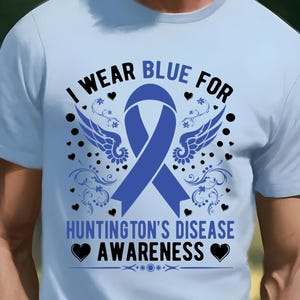 May include: Light blue t-shirt with the text "I WEAR BLUE FOR HUNTINGTON'S DISEASE AWARENESS". The design includes a blue ribbon, wings, hearts, and decorative flourishes. The shirt is a crew neck style.