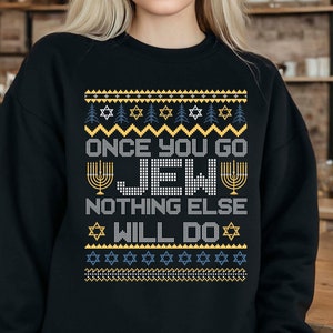 Puede incluir: Sudadera negra con el texto "ONCE YOU GO JEW NOTHING ELSE WILL DO" en una fuente pixelada. El diseño incluye estrellas de David y símbolos de menorá en amarillo, blanco y azul, que se asemejan a un suéter festivo.