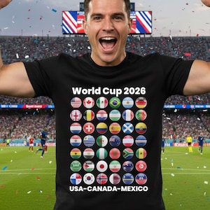 May include: A black t-shirt with the text "World Cup 2026" and a series of flag icons. The shirt also has the text "USA-CANADA-MEXICO" at the bottom. A man is wearing the shirt and celebrating with his arms raised in a stadium.