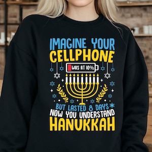 Op de afbeelding: Zwarte sweatshirt met een Hanukkah-thema ontwerp. Het ontwerp bevat de tekst "IMAGINE YOUR CELLPHONE WAS AT 10% BUT LASTED 8 DAYS NOW YOU UNDERSTAND HANUKKAH", een menora en Davidsterren.