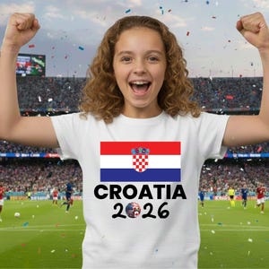 May include: A white t-shirt displaying the Croatian flag in red, white, and blue, with the text "CROATIA 2026" beneath. The image shows a person wearing the shirt, celebrating with arms raised, set against a stadium backdrop.