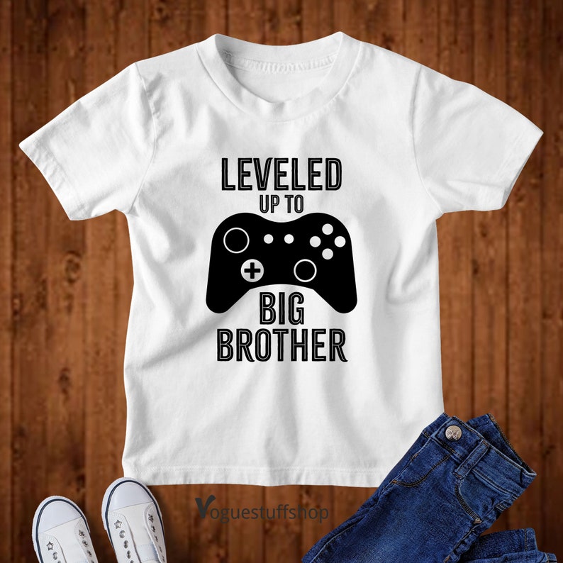 Może przedstawiać: Biała koszulka z czarnym tekstem i grafiką kontrolera do gier wideo, na kt&oacute;rej widnieje napis "Leveled Up To Big Brother".