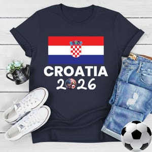 May include: Navy blue t-shirt with the Croatian flag and the text "CROATIA 2026". The design includes a soccer ball with a mixed flag design. The shirt is styled with denim shorts, white trainers, and a football.