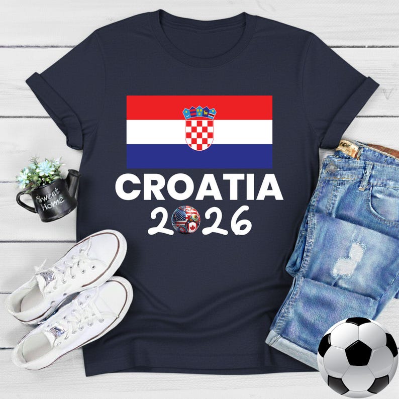 May include: Navy blue t-shirt with the Croatian flag and the text "CROATIA 2026". The design includes a soccer ball with a mixed flag design. The shirt is styled with denim shorts, white trainers, and a football.
