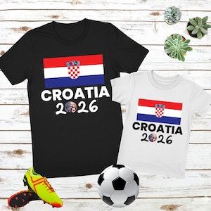 May include: Two t-shirts, one black and one white, featuring the Croatian flag and the text "CROATIA 2026". A football and a pair of neon green and black football boots are in the foreground. The shirts are on a light wood-grain surface.