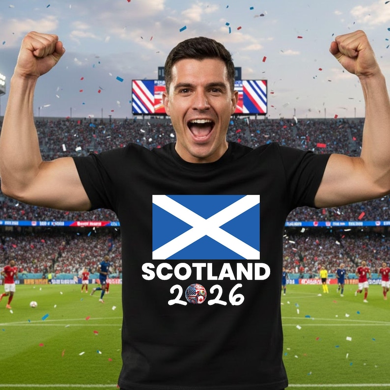 May include: Black t-shirt featuring the Scottish flag with a blue and white design. The text "SCOTLAND 2026" is printed below the flag. A person is wearing the shirt and celebrating with arms raised.