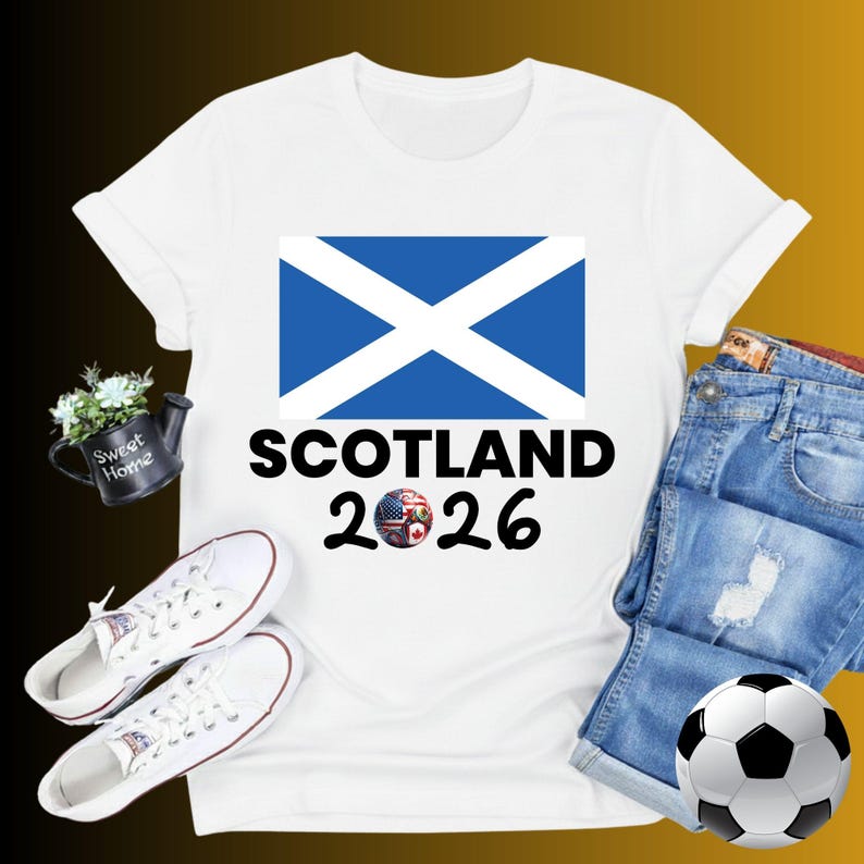 May include: White t-shirt with a blue and white Scottish flag design, the word "SCOTLAND" in black, and the year "2026". The design includes a soccer ball with flags. The shirt is paired with denim shorts, white sneakers, and a soccer ball.