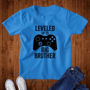 Może przedstawiać: Niebieska koszulka z czarnym nadrukiem kontrolera do gier wideo i tekstem "LEVELED UP TO BIG BROTHER".