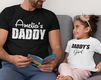 Puede incluir: Un padre y una hija con camisetas a juego. La camiseta del padre dice "Amelia's Daddy" y la camiseta de la hija dice "Daddy's Girl".