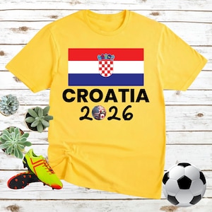 May include: A yellow t-shirt with the Croatian flag and the text "CROATIA 2026". A soccer ball with flags of various countries is below the text. A football and football boots are also in the image.