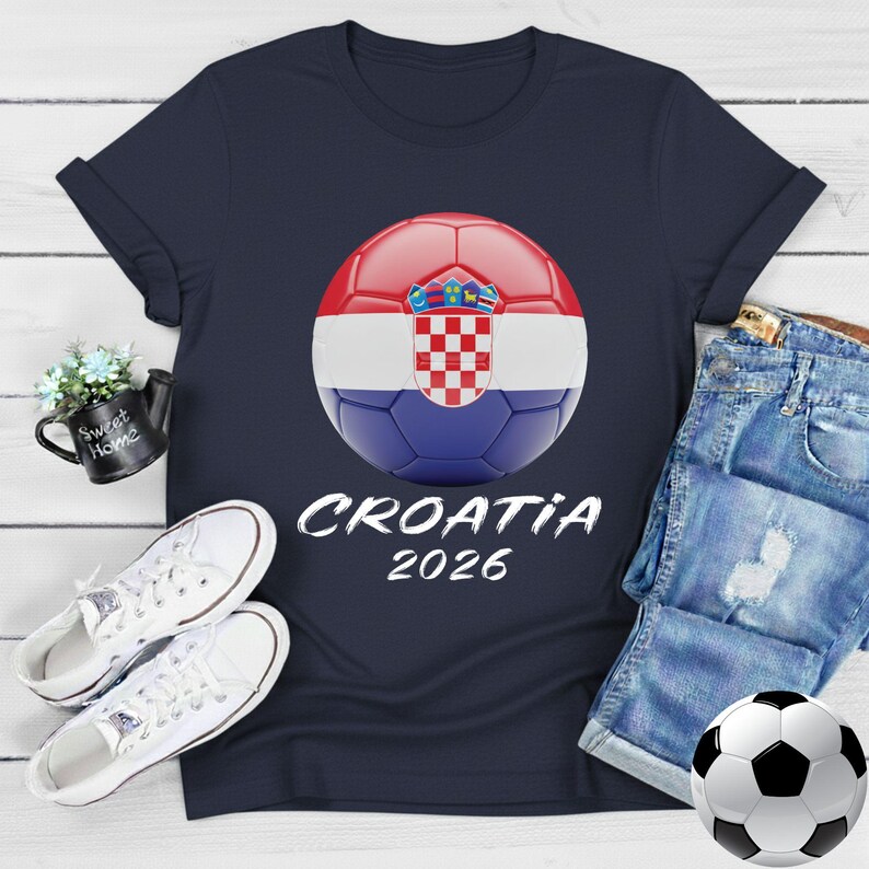 K&ouml;nnte beinhalten: Marineblaues T-Shirt mit einem Fu&szlig;ball-Design in den Farben der kroatischen Flagge und dem Text "CROATIA 2026". Das Bild zeigt wei&szlig;e Turnschuhe und Jeansshorts, was einen l&auml;ssigen, sportlichen Look erzeugt.